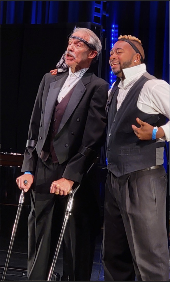Aarron Loggins and Scott Sedar acting on stage. One white gentleman with crutches wears a tie around his head and a Black man stands next to him with his arm around him. Both are in black slacks, button up shirts, and vests. The white man wears a suit jacket also.
