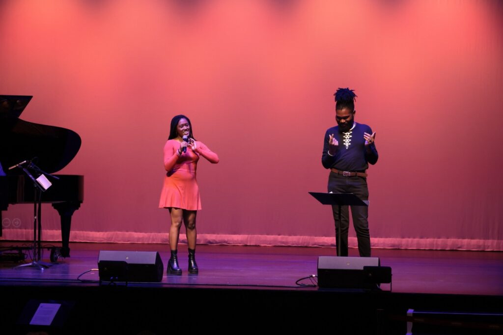 A Black woman in a red dress and black boots speaks on stage. A Black man with black hair pulled on top of his head, a black shirt and gold necklace signs.