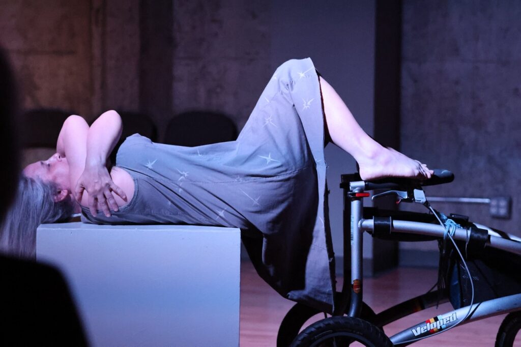 A white woman hugging herself is wearing a gray dress lays on her back on a gray cube and her feet propped up on her wheel chair. She has long gray hair draped back over the cube.
