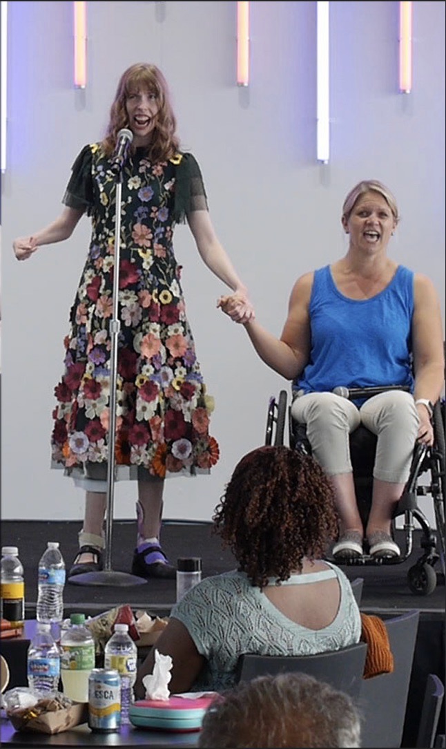 Two Women Singing and holding hands - One woman standing in a floral dress with shoulder length red hair - One woman in a wheel chair has blonde hair and wears a blue shirt and white capris. And audience looks on