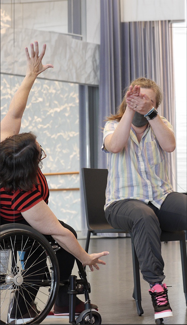 Suzanne Richard and Corina Dalzell are demonstrating framing in dance - One woman in a wheel chair reaching in a red and black shirt, another woman sitting in a chair in a pastel striped shirt clapping.