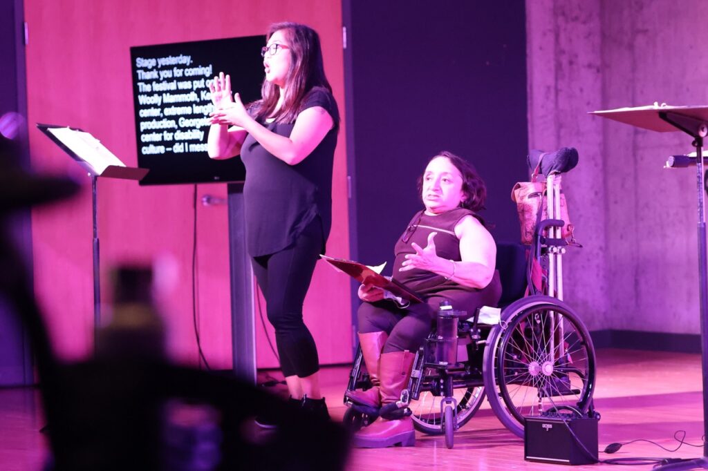 Two women on stage, one in a wheelchair with brown bobbed hair speaking and one standing with long black hair signing to introduce the Open Doors Festival. Both women are wearing black tops and bottoms.